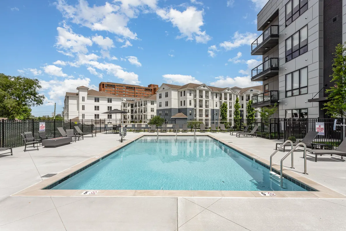 An exterior shot of a swimming pool surrounded by lounge chairs, adjacent to modern residential buildings and framed by trees. The setting is perfect for both relaxation and social gatherings, with space for sunbathing and enjoying the outdoors.