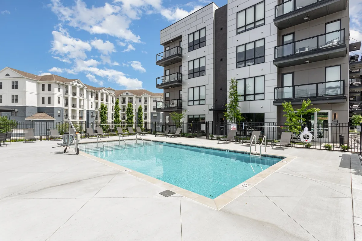 This image showcases a spacious swimming pool surrounded by the balconies of modern apartment buildings and lush green landscaping. Lounge chairs line the poolside, creating an inviting space for socializing, swimming, or relaxing. The communal atmosphere is evident, with areas for both sunbathing and gathering.