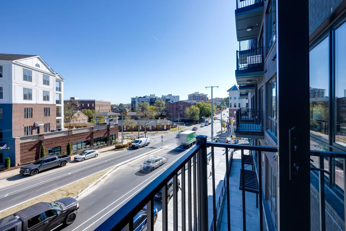 An exterior shot from a balcony looking onto a lively street scene, with railings in the foreground and surrounding buildings visible. The atmosphere captures the urban pulse and walkability of the neighborhood.