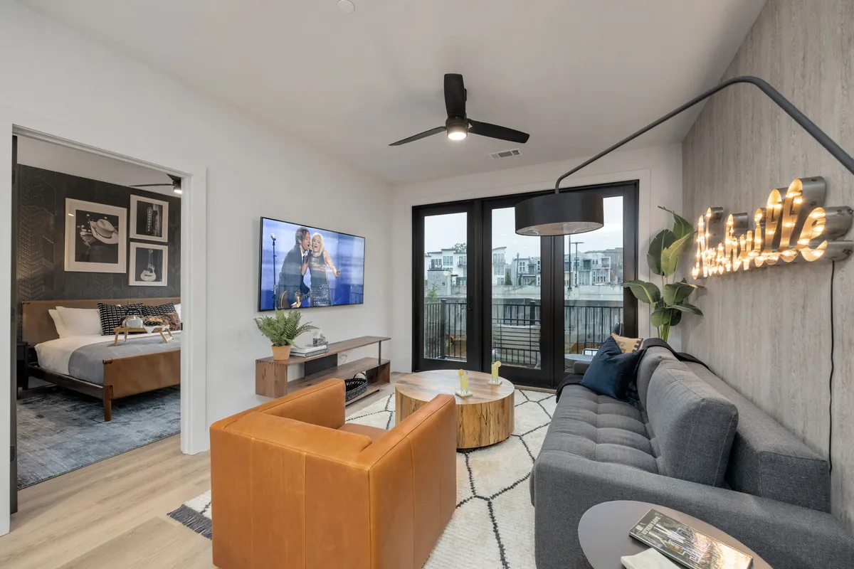 The image showcases a bright, contemporary living area furnished with a plush sofa, stylish coffee table, and a large TV. A distinctive 'Nashville' sign adds local flair, while expansive windows bathe the space in natural light and reveal a private balcony, perfect for enjoying sunsets or city views. The overall atmosphere is inviting and ideal for relaxation, socializing, and gathering with friends or family.