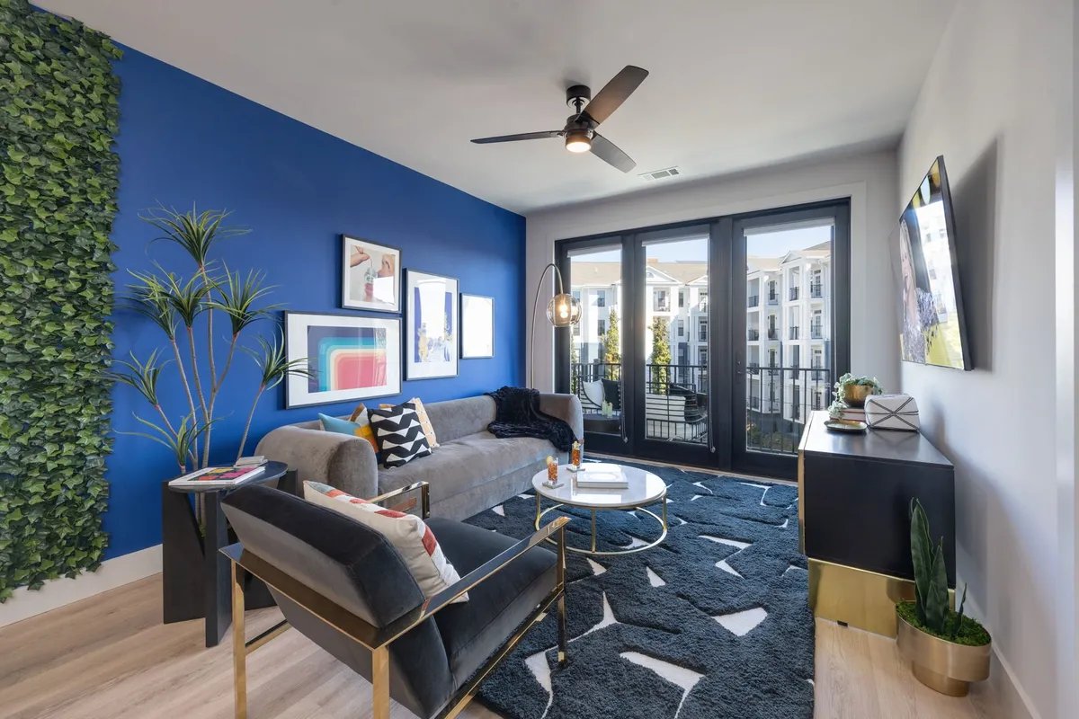 This image showcases a stylish living area with a blue accent wall, greenery, a comfortable sofa, artwork, a coffee table, and large windows that open onto a balcony. The decor is contemporary and inviting, with a layout that supports both group gatherings and quieter moments.