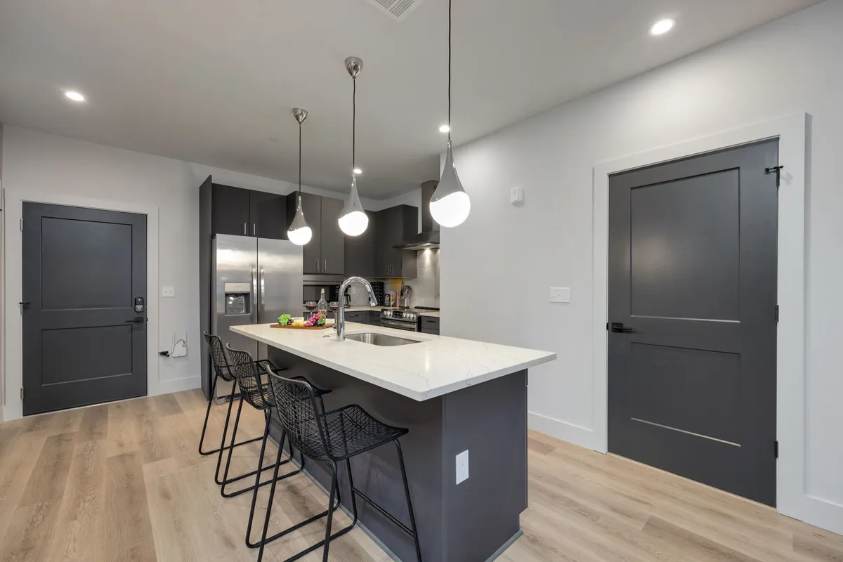 A modern kitchen scene with a large island, multiple bar stools, pendant lights, and sleek stainless steel appliances. The arrangement suggests a warm, inviting space designed for entertaining and shared meals, with a contemporary yet welcoming atmosphere.