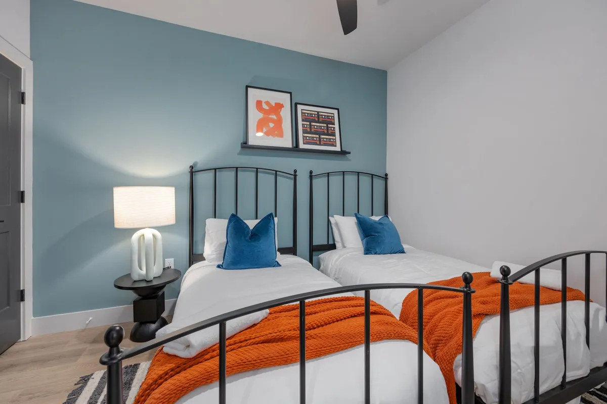 A playful and comfortable bedroom featuring two twin beds with distinctive black metal frames, orange throw blankets, blue accent pillows, and artwork on the wall. The room feels flexible and ideal for families or groups.