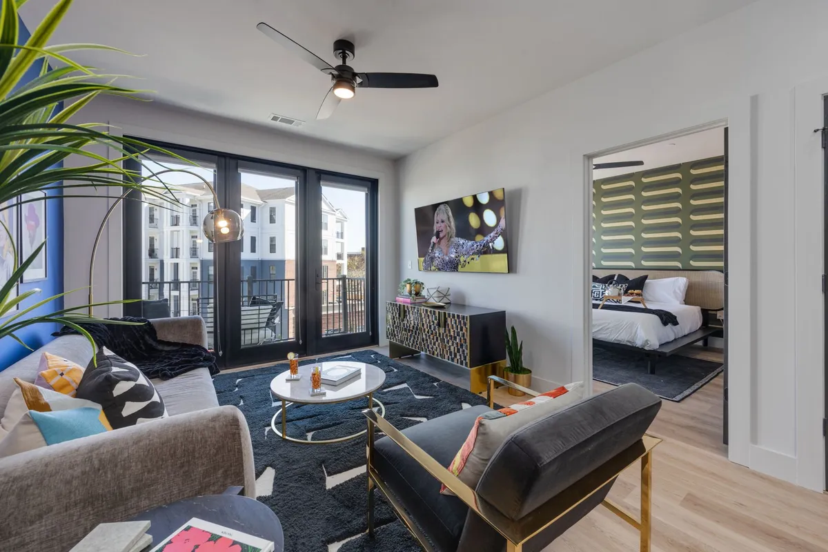 This image features an open, vibrant living space with large windows opening to a balcony view, a stylish coffee table, an artfully arranged couch and chairs, and a television displaying lively content. The room exudes both comfort and sociability, with ample seating for groups and visual access to the balcony—a key feature in the section.