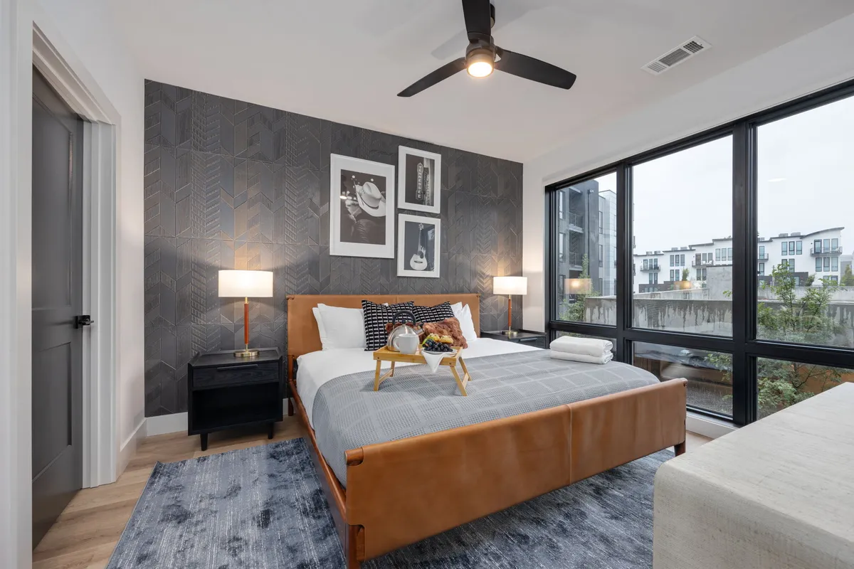 A serene bedroom with a leather-framed bed, large windows allowing natural light to filter in, decorative wall art, and stylish nightstands with lamps. The space feels modern, inviting, and perfect for unwinding.