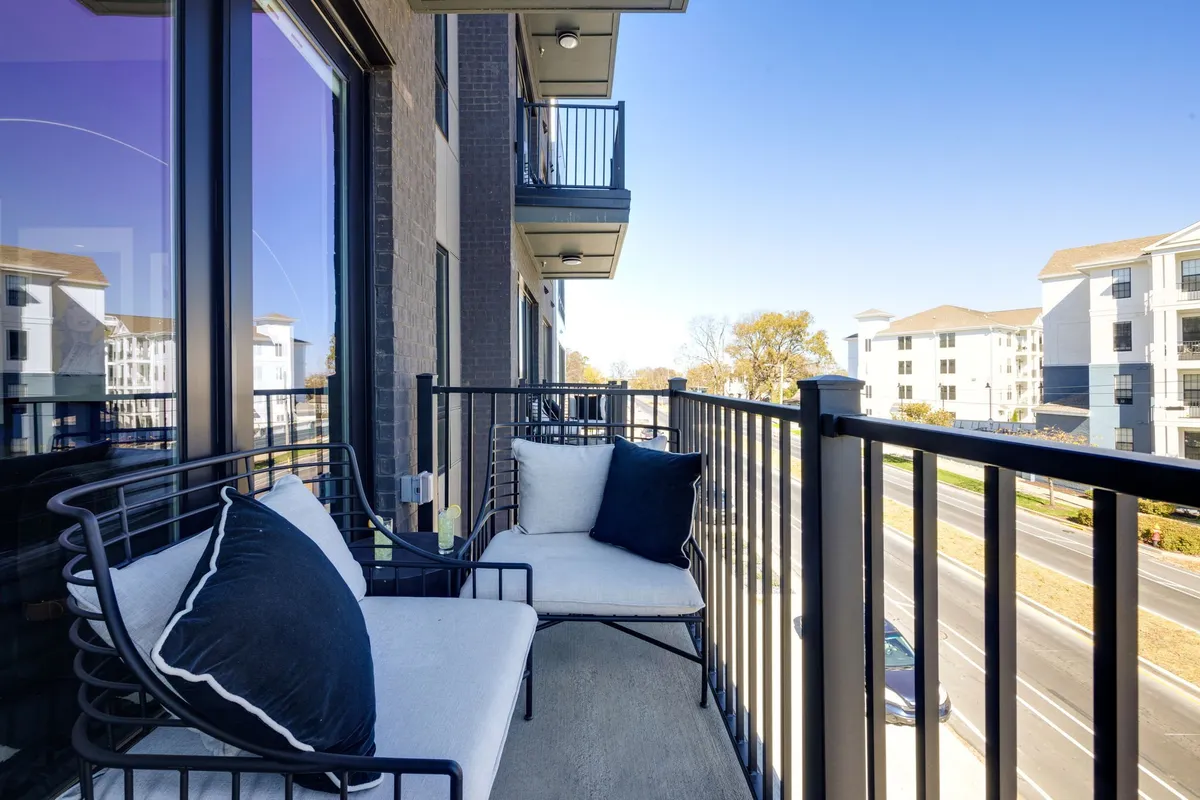 A private balcony with comfortable seating offers a view of the street below, surrounded by railings and bathed in natural light. The setup is perfect for quiet relaxation, morning coffee, or socializing in a peaceful outdoor environment.